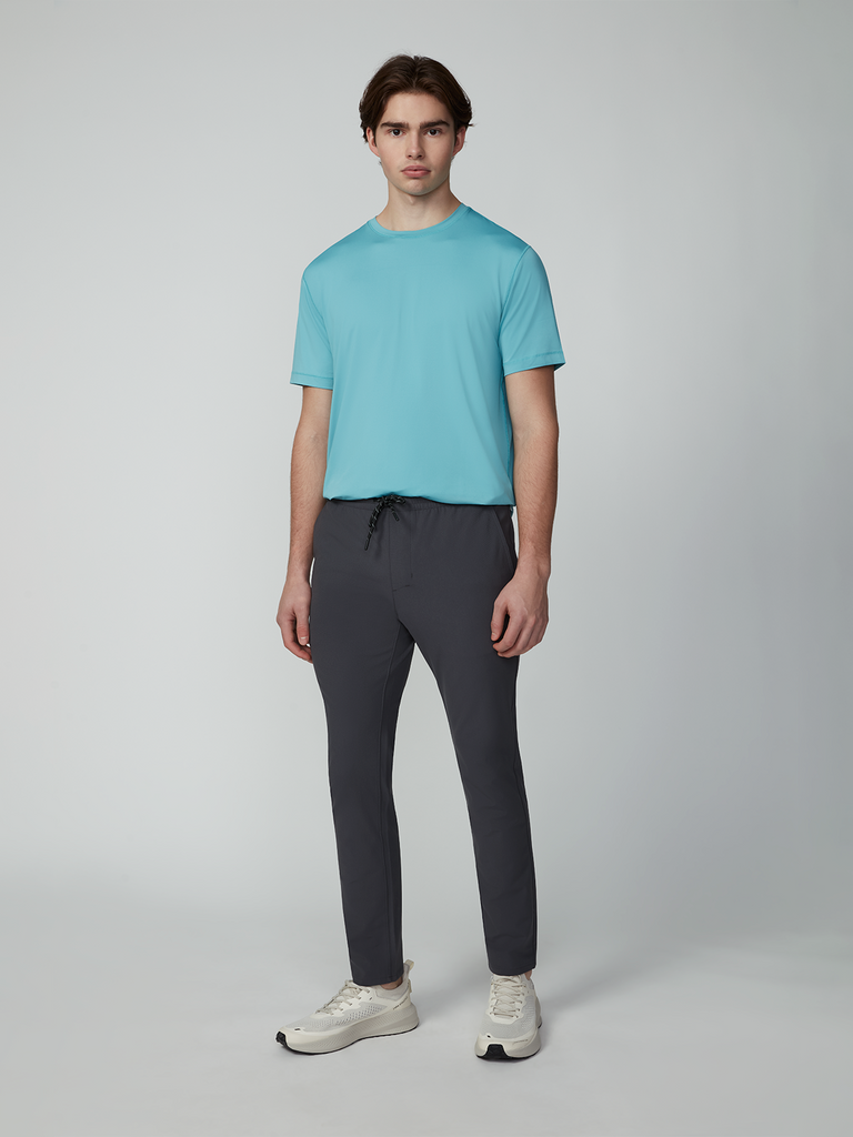 A man stands against a plain light gray background, wearing a light blue short-sleeve t-shirt and TECH APPAREL MENS FLUX 2.0 PULL-ON PANT in dark gray with a drawcord waist, plus light sneakers. He faces forward with a neutral expression.