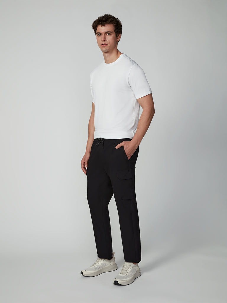 A man stands against a light gray background, wearing a white tee, white sneakers, and TECH APPARELs MENS FLUX 2.0 PULL-ON CARGO PANT. He has one hand in his pocket and looks slightly to the side.