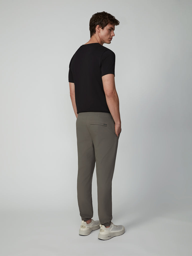 A man faces away, wearing TECH APPAREL’s MENS DRI-FLEX JOGGER in gray, a black short-sleeve shirt, and white sneakers against a plain light gray background.