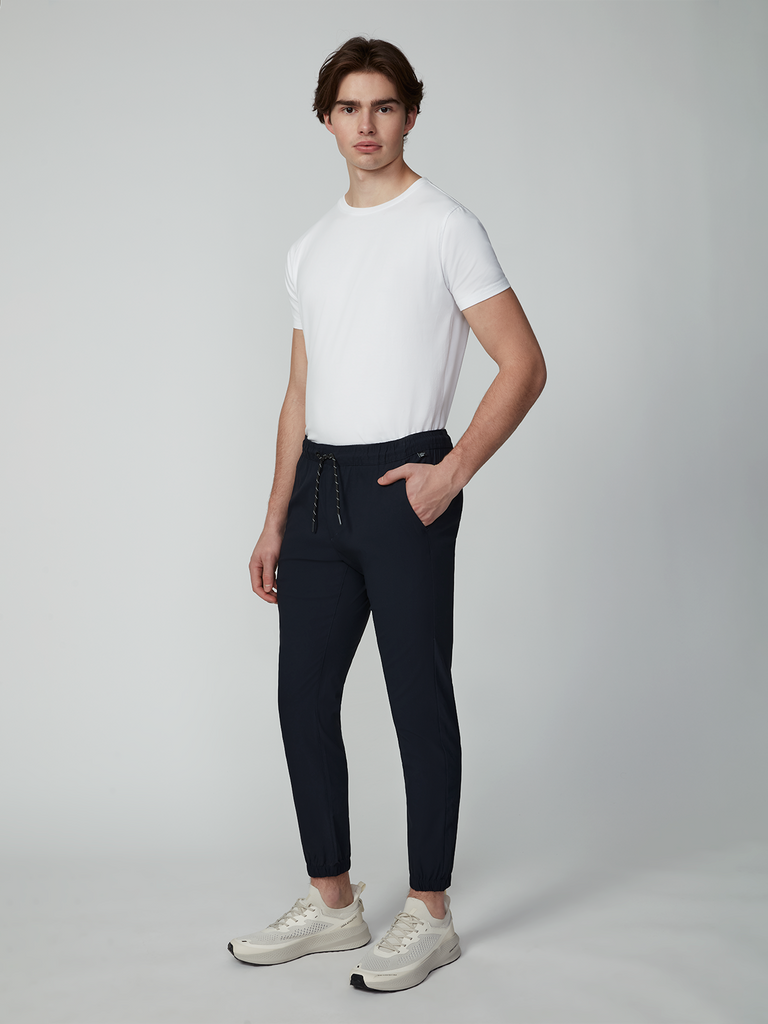 A young man with short brown hair and a neutral expression stands against a plain background, wearing TECH APPAREL MENS DRI-FLEX JOGGER with an athletic fit and drawstrings, paired with a white t-shirt and white sneakers.