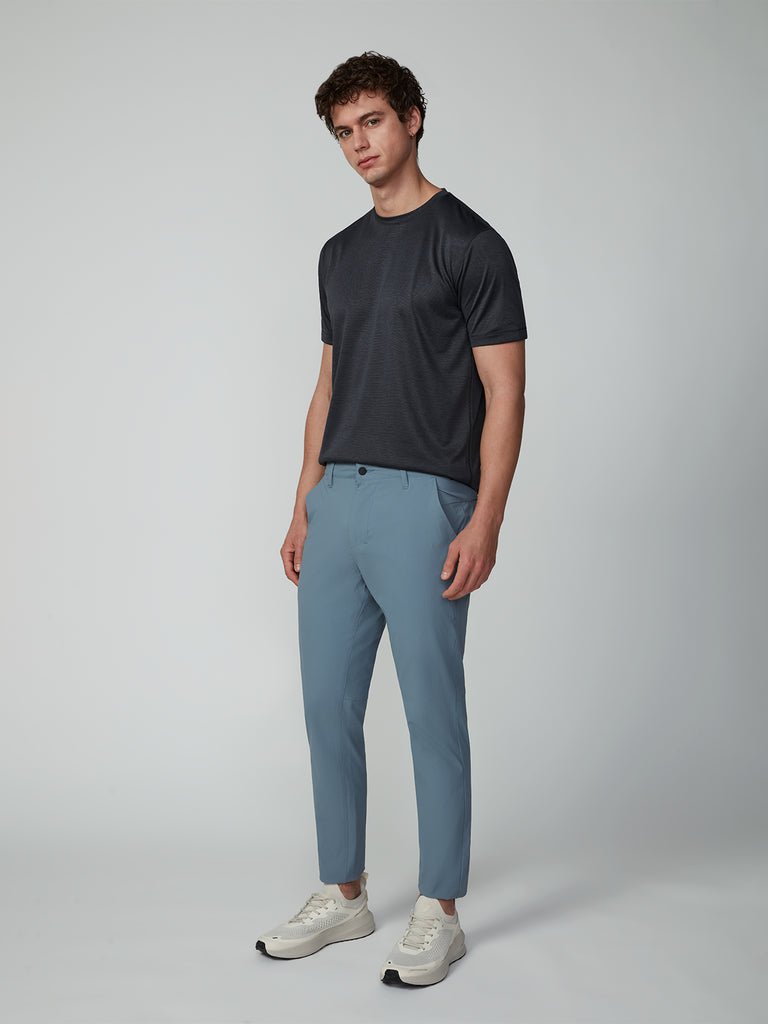 A man stands against a light gray background, wearing a dark gray short-sleeve t-shirt, TECH APPAREL MENS DRI-FLEX PANTS in light blue, and white sneakers. He faces slightly left with a neutral expression and relaxed posture.