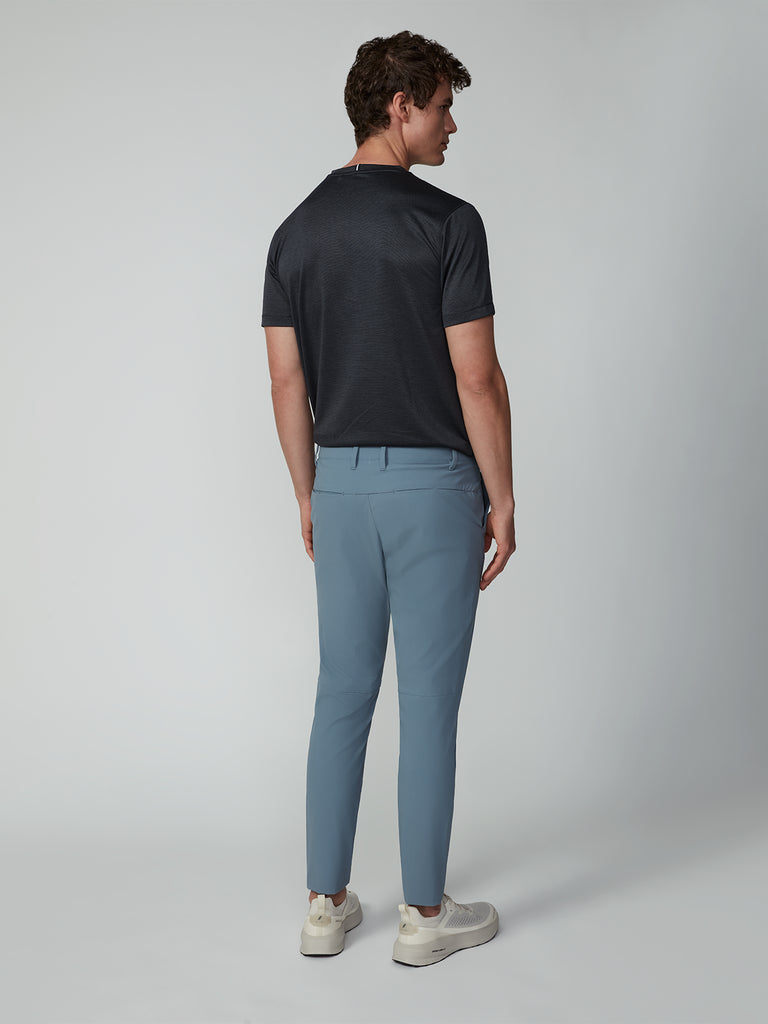 A man faces away from the camera in a dark short-sleeve shirt, TECH APPAREL MENS DRI-FLEX PANTS in wrinkle-resistant light blue, and white sneakers, standing against a plain light gray background.