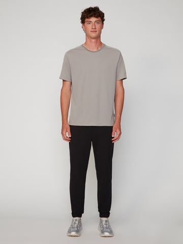 A young man with short, curly brown hair stands facing forward in a light gray tee, TECH APPAREL MEN’S 360 KNIT CLASSIC FIT PULL-ON JOGGER pants, and gray sneakers against a plain background. His arms rest at his sides.