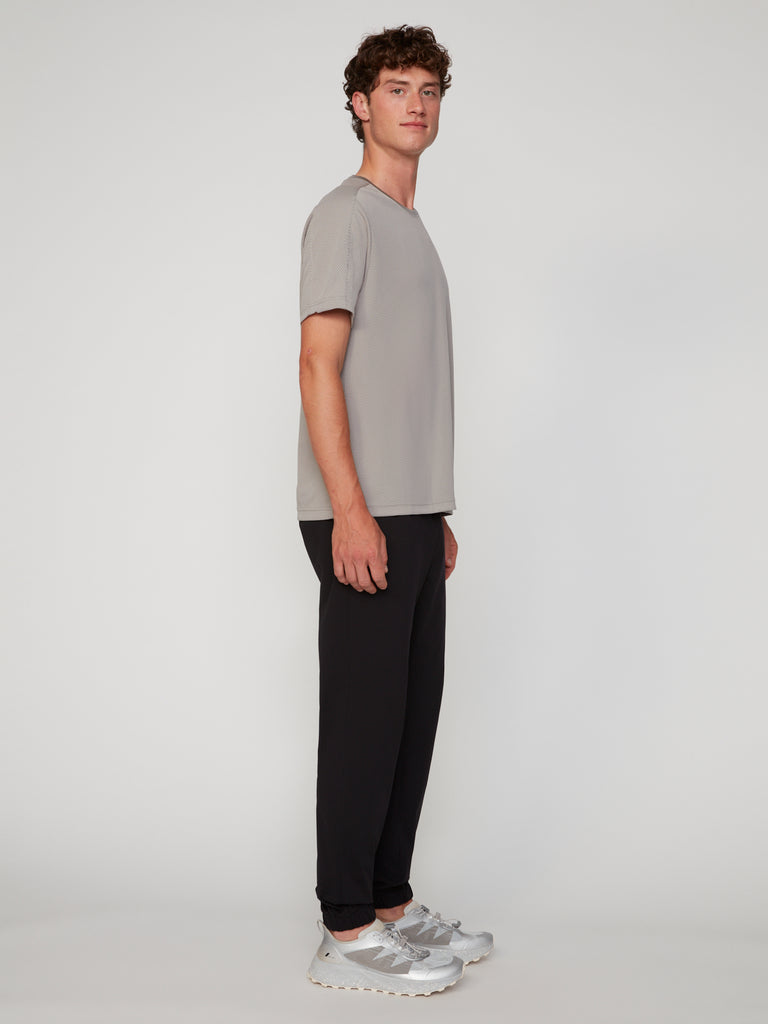 A man stands in profile against a plain background, wearing a light gray short-sleeve t-shirt, white sneakers, and black TECH APPAREL MEN’S 360 KNIT CLASSIC FIT PULL-ON JOGGER pants.