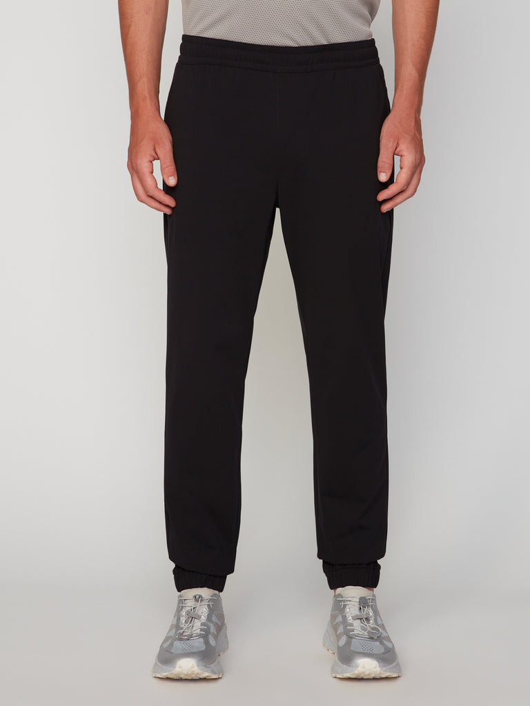 Wearing TECH APPAREL’s MEN’S 360 KNIT CLASSIC FIT PULL-ON JOGGER in black and gray athletic shoes, a person stands against a white background with hands at their sides; only the lower half of their body is shown.