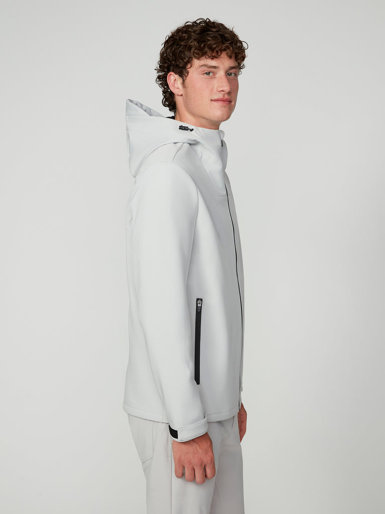 A young man with curly hair stands sideways against a white background, wearing TECH APPARELs MENS BONDED FLEECE JACKET in light gray with black zippers and matching pants. He faces the camera with a slight smile.