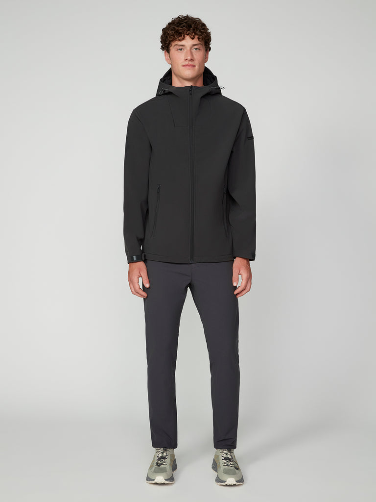 A young man with curly hair stands facing forward, wearing a black TECH APPAREL MENS BONDED FLEECE JACKET, dark pants, and gray sneakers against a plain light gray background.