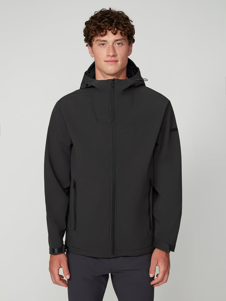 A young man with curly brown hair wears a TECH APPAREL MENS BONDED FLEECE JACKET in black with a hood and zip front paired with dark pants, standing against a light gray background and facing the camera with a neutral expression.