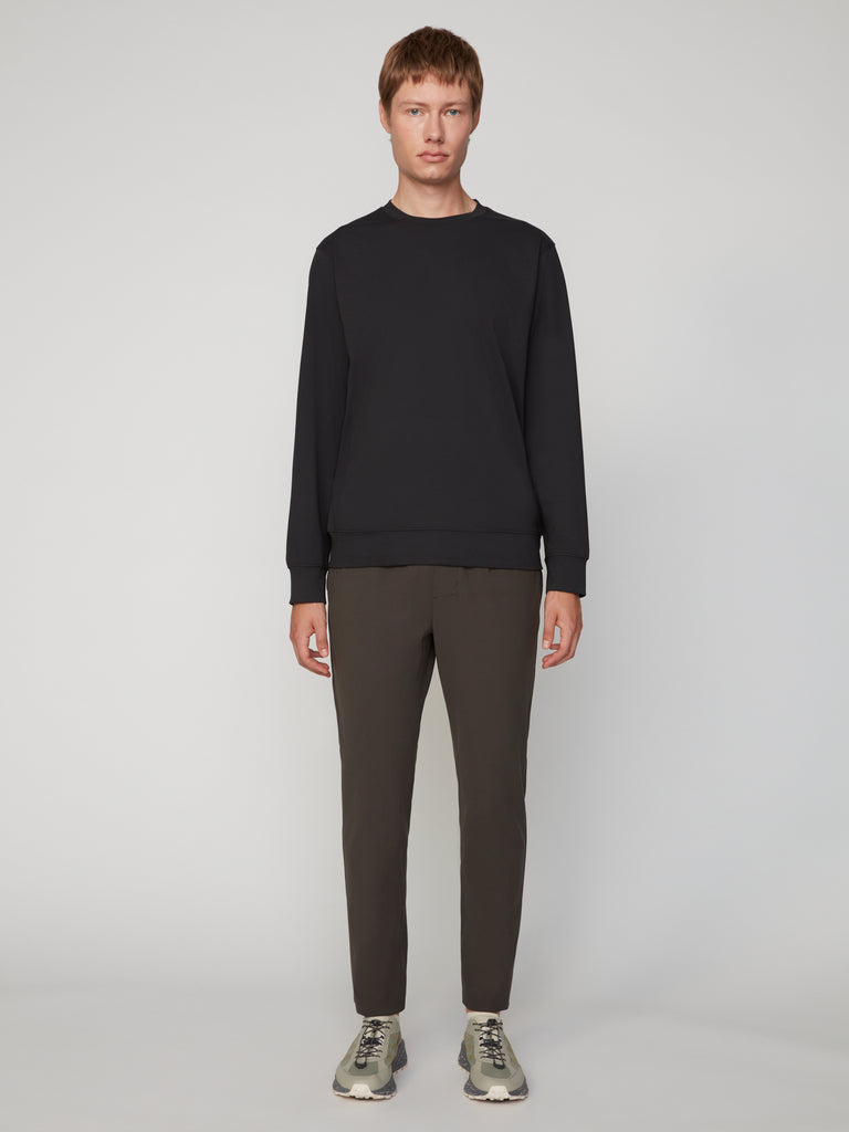 A man stands facing forward against a plain light gray background, wearing a black long-sleeve sweatshirt, TECH APPAREL MEN’S FLUX 2.0 CLASSIC FIT PULL-ON CHINO PANT in dark olive, and gray sneakers.