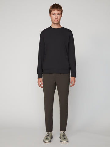 A man stands facing forward against a plain light gray background, wearing a black long-sleeve sweatshirt, TECH APPAREL MEN’S FLUX 2.0 CLASSIC FIT PULL-ON CHINO PANT in dark olive, and gray sneakers.