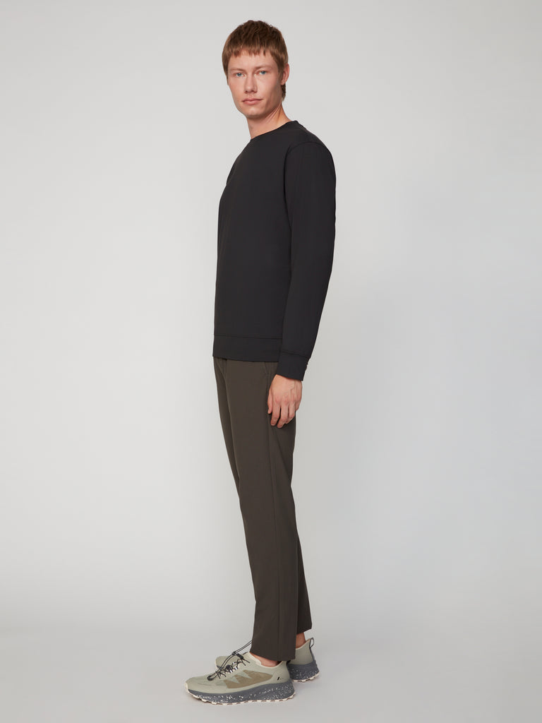 A man stands sideways, looking at the camera, wearing a black long-sleeve top, TECH APPAREL MEN’S FLUX 2.0 CLASSIC FIT PULL-ON CHINO PANT, and light gray chunky sole sneakers against a plain light background.