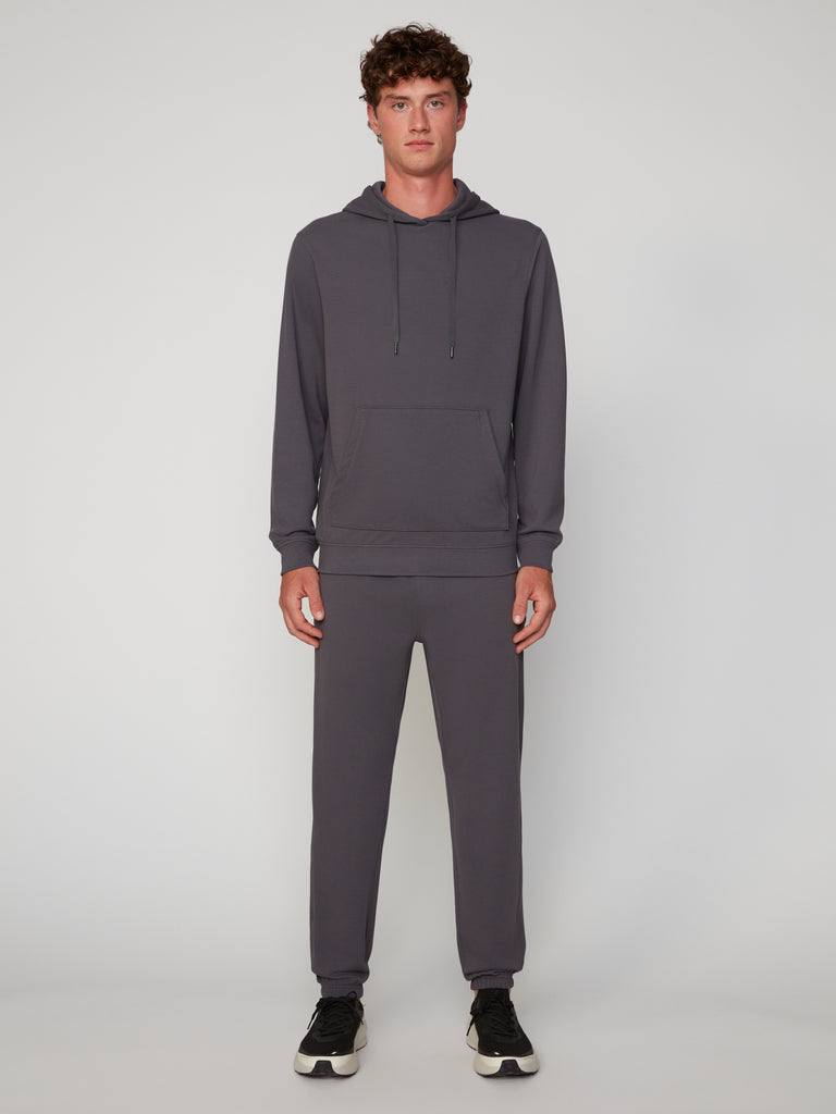 A young man faces forward wearing TECH APPAREL’s MEN’S MICRO:WAFFLE CLASSIC FIT PULL-ON CHINO PANT in charcoal gray with black sneakers, set against a plain light gray background.