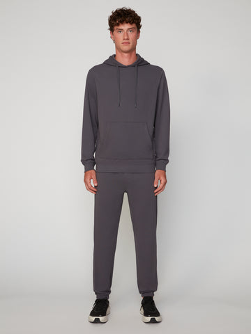 A young man faces forward wearing TECH APPAREL’s MEN’S MICRO:WAFFLE CLASSIC FIT PULL-ON CHINO PANT in charcoal gray with black sneakers, set against a plain light gray background.
