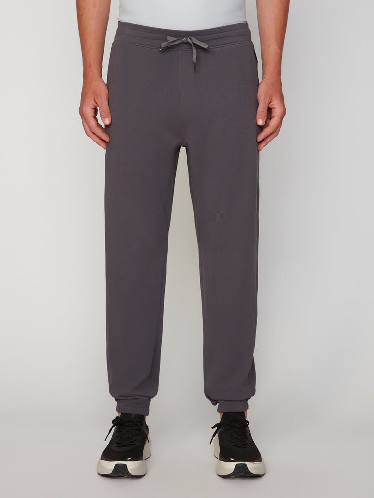 A person is shown from the waist down in TECH APPAREL MEN’S MICRO:WAFFLE CLASSIC FIT PULL-ON CHINO PANT in dark gray, paired with black and white sneakers, standing against a plain white background. Only the lower torso and legs are visible.