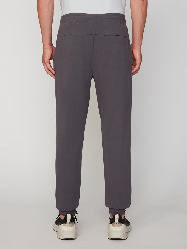 A person is shown from the back, standing against a white background, wearing TECH APPAREL MEN’S MICRO:WAFFLE CLASSIC FIT PULL-ON CHINO PANT in gray with white sneakers.