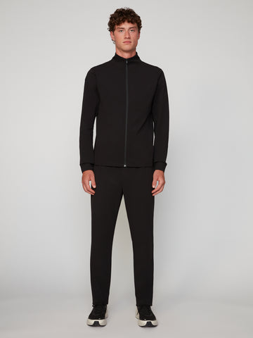 A man stands facing forward against a plain light background, wearing a black zip-up jacket, TECH APPAREL MEN’S FLEXURA RELAXED FIT CHINO PANT, and black sneakers with white soles.