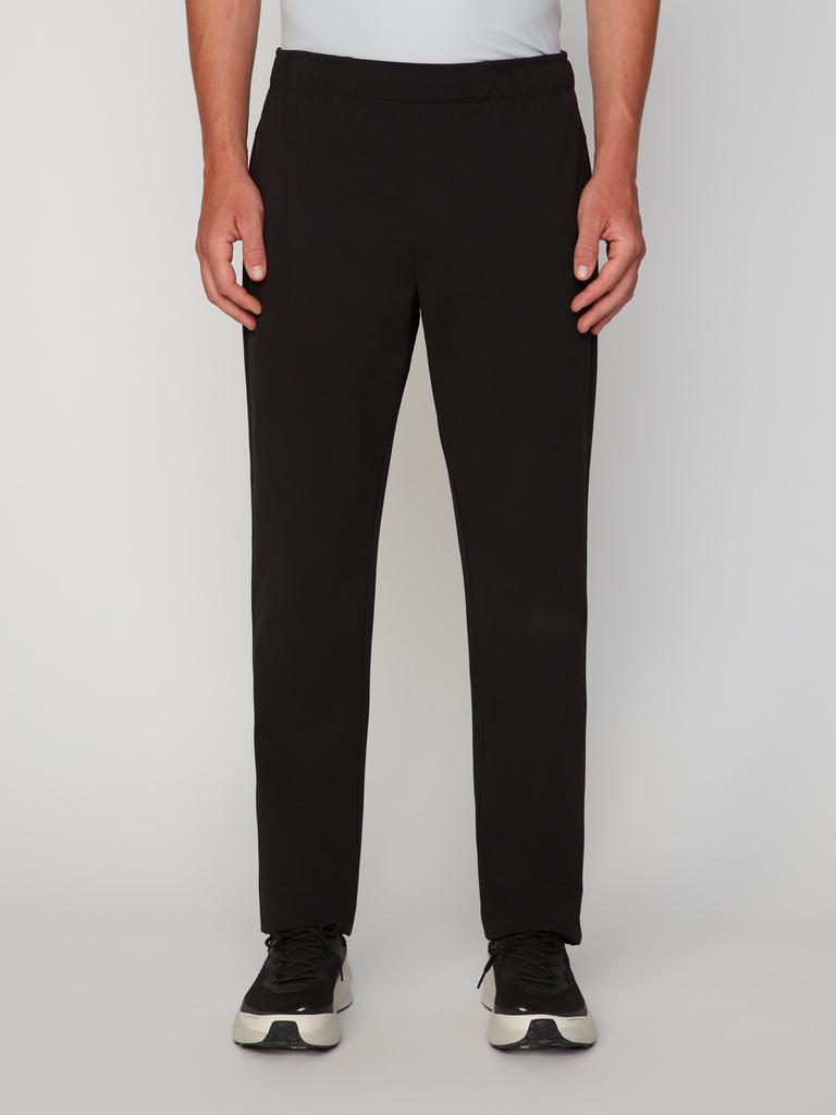 A person wears black TECH APPAREL MEN’S FLEXURA RELAXED FIT CHINO PANT and black-and-white sneakers, standing against a plain light background. Only the lower torso and legs are visible.