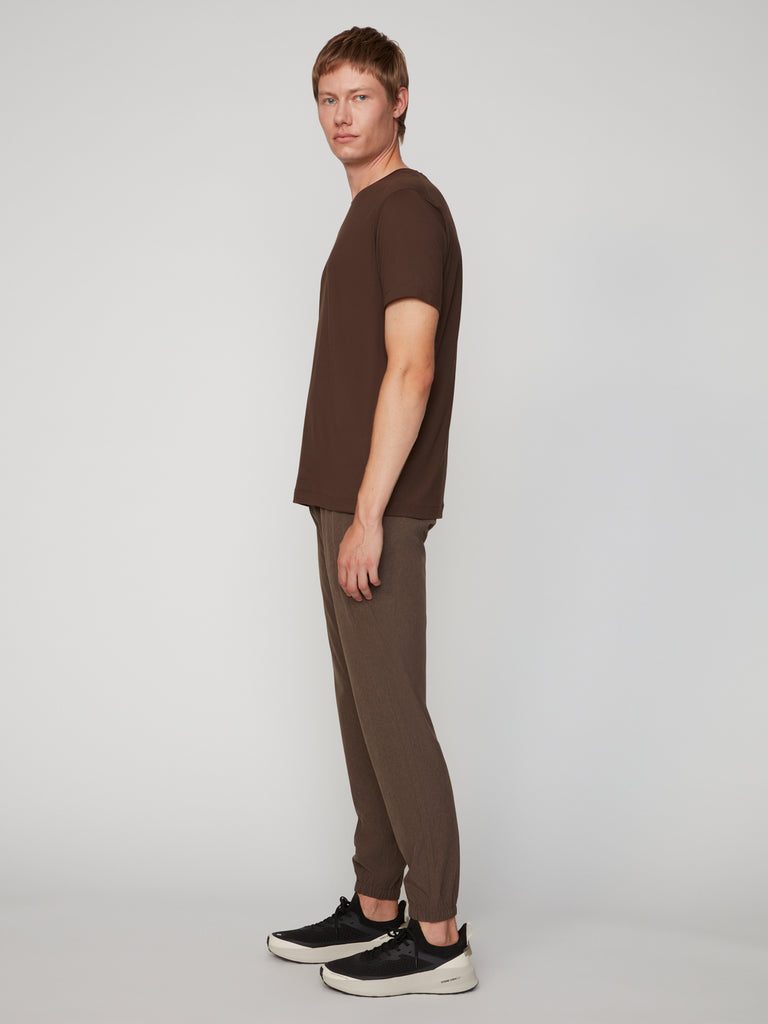 A young man stands sideways against a plain background, wearing TECH APPAREL’s MEN’S 360:FLEX CLASSIC FIT PULL-ON CHINO JOGGER, styled with a brown T-shirt and black-and-white sneakers.