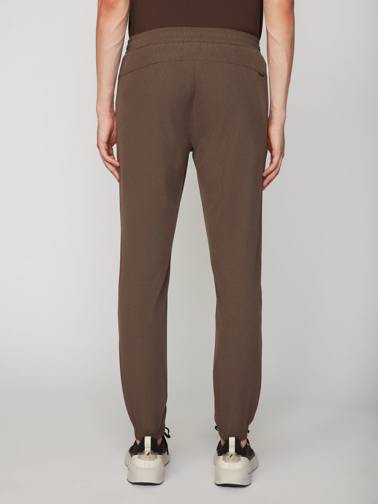 A person faces away from the camera, wearing TECH APPAREL MEN’S 360:FLEX CLASSIC FIT PULL-ON CHINO JOGGER pants in brown with a pull-on waistband and white sneakers, set against a plain light gray background.