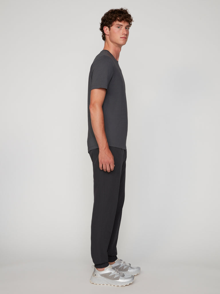 A young man with curly hair stands in profile against a plain background, wearing a dark grey t-shirt, TECH APPAREL MEN’S 360:FLEX CLASSIC FIT PULL-ON CHINO JOGGER, and white sneakers.