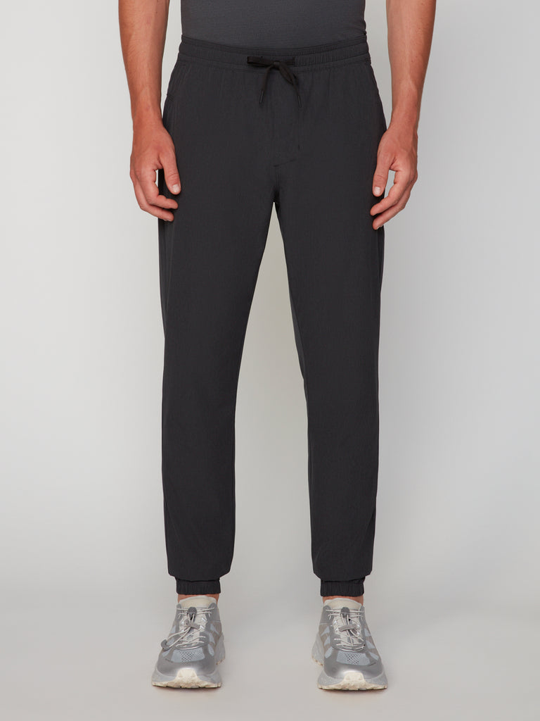 A person wears TECH APPAREL MEN’S 360:FLEX CLASSIC FIT PULL-ON CHINO JOGGER in black and gray sneakers, shown from the waist down against a plain, light background.