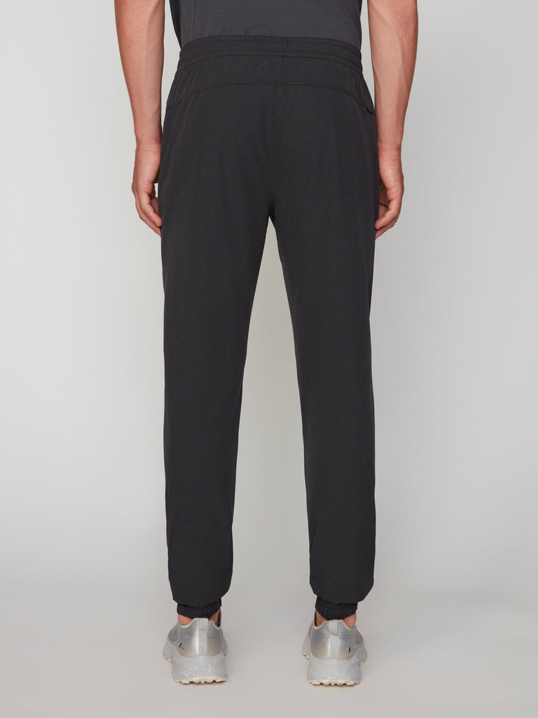 A person wearing TECH APPAREL MEN’S 360:FLEX CLASSIC FIT PULL-ON CHINO JOGGER with an elastic waistband and cuffed ankles stands facing away from the camera, paired with silver sneakers against a plain light background.