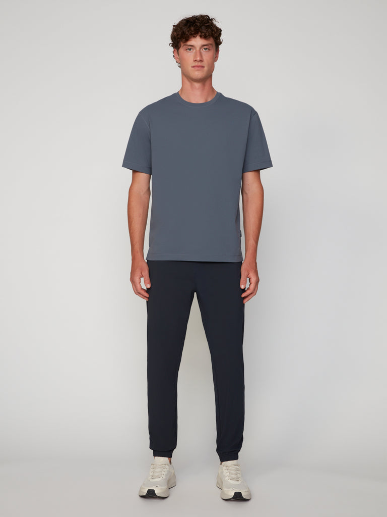 A young man stands facing forward against a plain light background, wearing a slate blue short-sleeve tee, white sneakers, and TECH APPAREL MEN’S FLEXLITE CLASSIC FIT PULL-ON JOGGER pants.