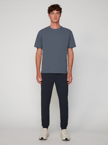 A young man stands facing forward against a plain light background, wearing a slate blue short-sleeve tee, white sneakers, and TECH APPAREL MEN’S FLEXLITE CLASSIC FIT PULL-ON JOGGER pants.