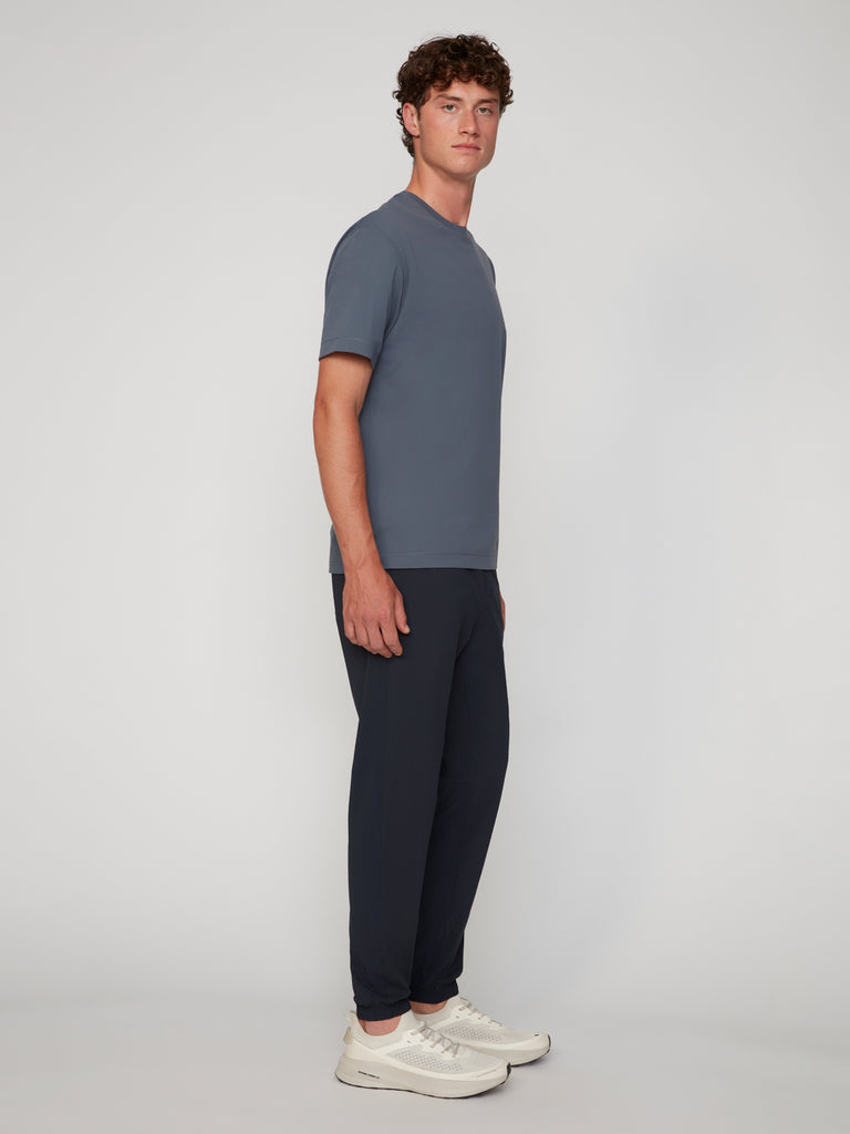 A man with curly hair models the TECH APPAREL MEN’S FLEXLITE CLASSIC FIT PULL-ON JOGGER, paired with a blue-gray tee and white sneakers, highlighting its lightweight FlexLite performance fabric against a plain backdrop.
