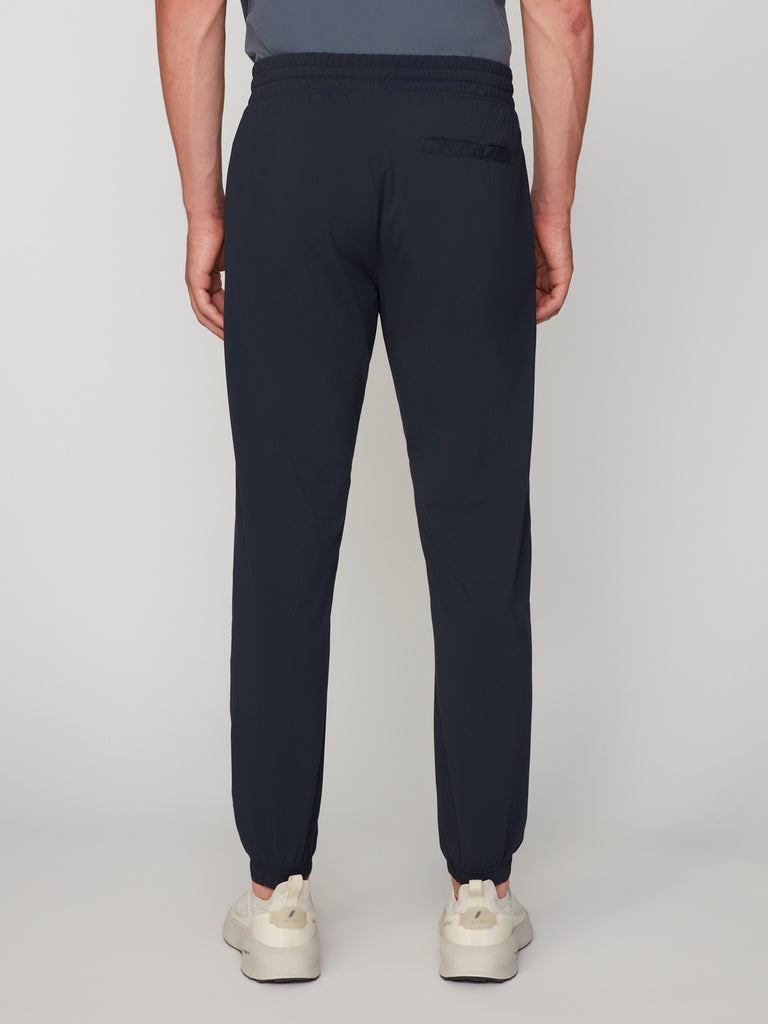 A man faces away, wearing TECH APPAREL’s MEN’S FLEXLITE CLASSIC FIT PULL-ON JOGGER in dark navy with an elastic waistband, cuffs, single right back pocket, and white sneakers. The FlexLite fabric ensures lightweight performance.