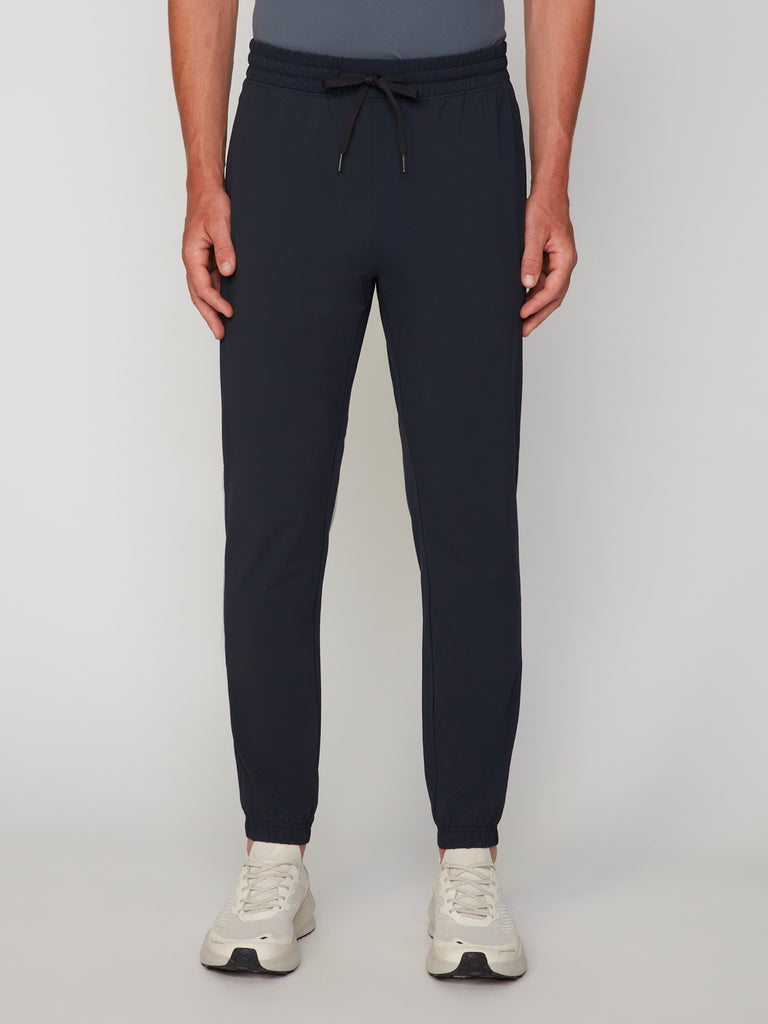 A person wears TECH APPAREL MEN’S FLEXLITE CLASSIC FIT PULL-ON JOGGER pants in dark navy, featuring FlexLite fabric, an elastic waistband, and drawstring. Paired with light sneakers, only the lower body is visible as the image is cropped at the shoulders.