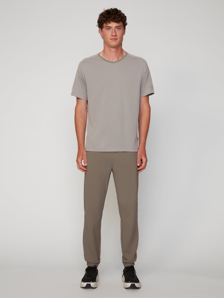 A person stands facing forward against a light-gray background, wearing a light gray t-shirt, TECH APPAREL MEN’S FLEXLITE CLASSIC FIT PULL-ON JOGGER in taupe, and black sneakers with white soles.