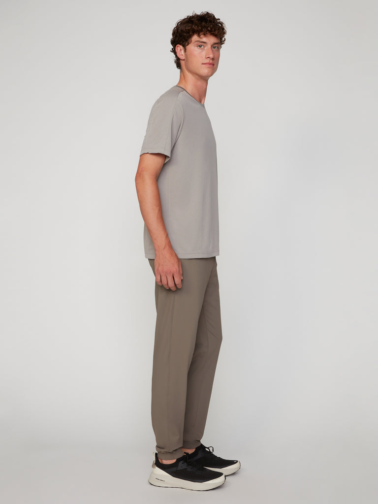 A young man with curly hair stands in profile against a plain light background, wearing a light gray t-shirt, TECH APPAREL MEN’S FLEXLITE CLASSIC FIT PULL-ON JOGGER, and black sneakers with white soles.
