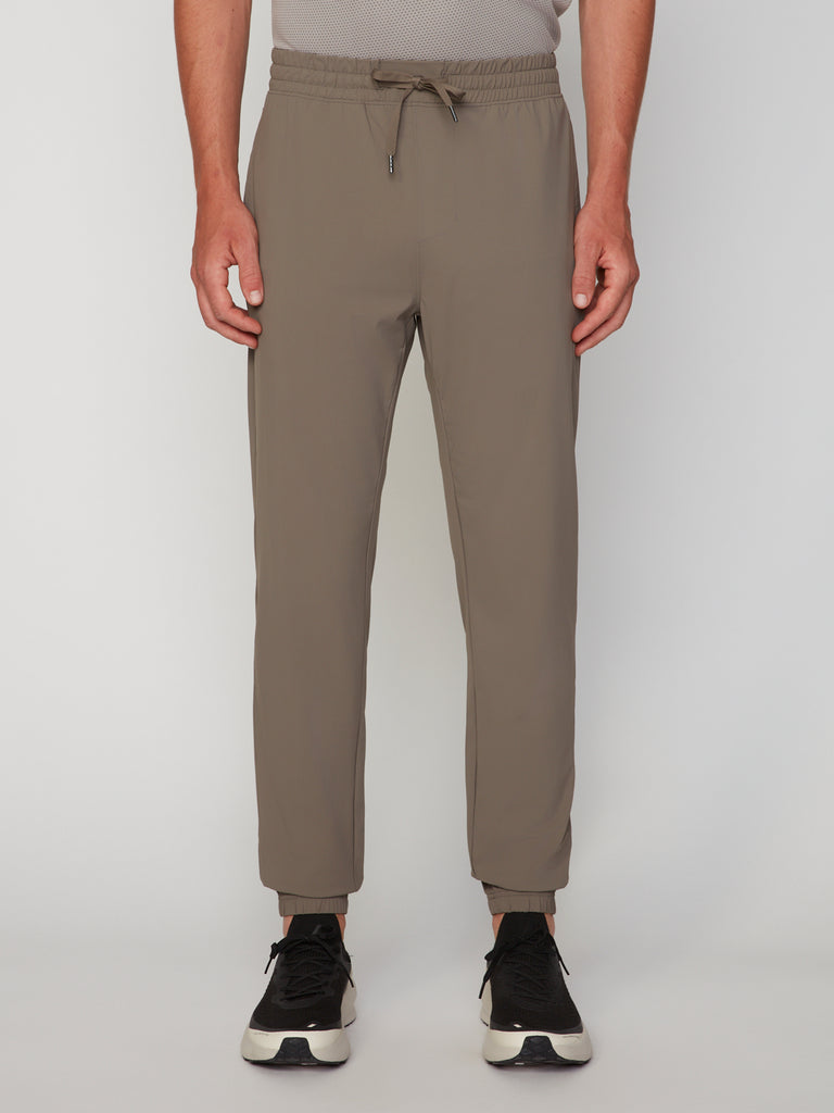 A man wears TECH APPAREL’s MEN’S FLEXLITE CLASSIC FIT PULL-ON JOGGER in taupe with black and white sneakers, standing against a plain light background. The image is cropped at the chest.