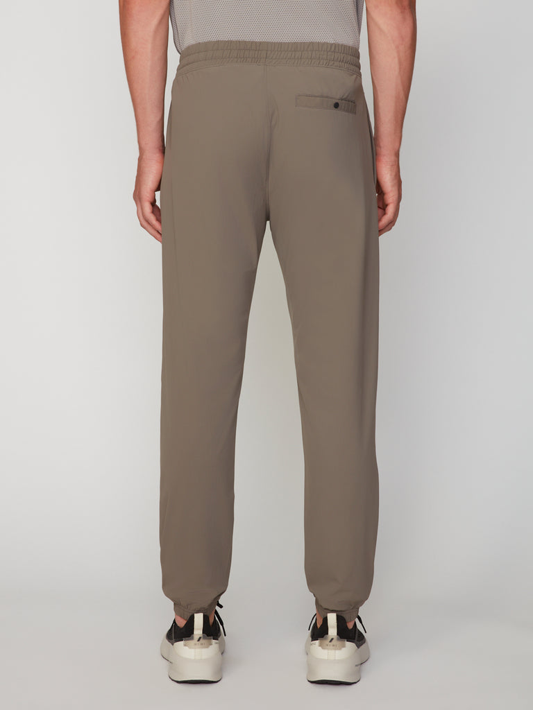 A person faces away, wearing TECH APPAREL’s MEN’S FLEXLITE CLASSIC FIT PULL-ON JOGGER in taupe with an elastic waistband and one back pocket, styled with white sneakers against a plain, light background.