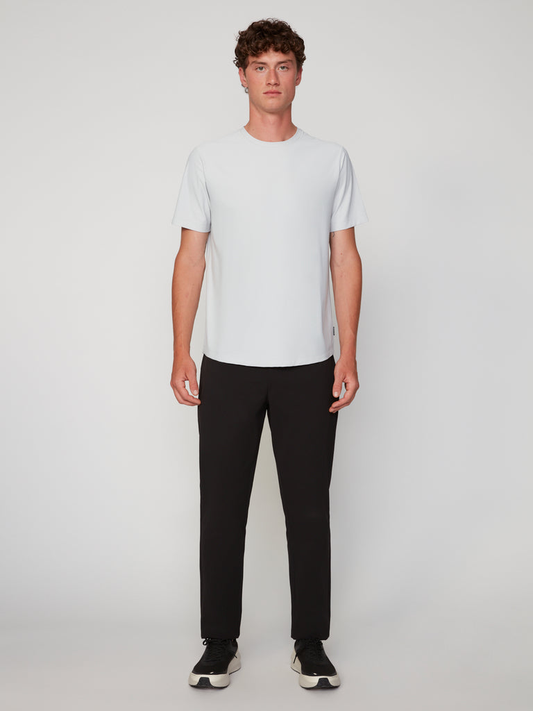 A young man with curly hair faces forward against a plain background, wearing the TECH APPAREL MEN’S MINI WAFER CLASSIC FIT T-SHIRT for breathable comfort, paired with black pants and sneakers. He has a neutral expression and relaxed posture.
