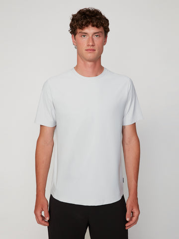 A man with short curly brown hair, wearing black pants and facing the camera neutrally, models the TECH APPAREL MEN’S MINI WAFER CLASSIC FIT T-SHIRT made from breathable, moisture-managing fabric against a plain light background.