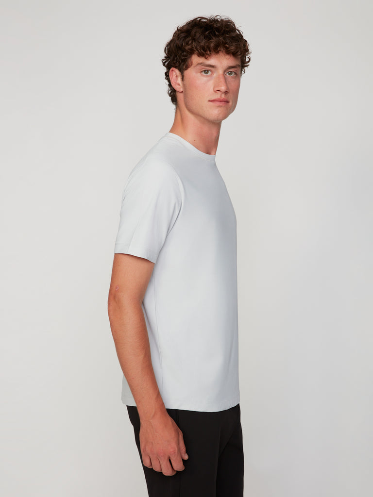 A young man with curly brown hair, facing left, wears the TECH APPAREL MEN’S MINI WAFER CLASSIC FIT T-SHIRT—made from breathable Mini Wafer fabric for moisture management—and black pants against a plain light background.