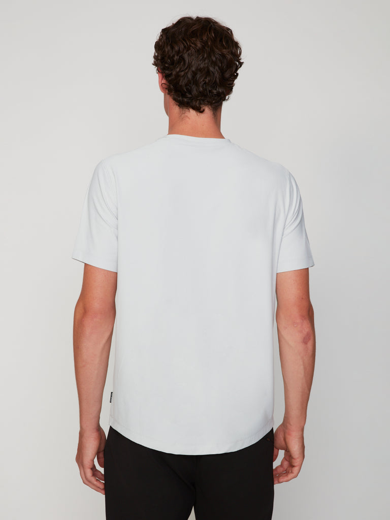 A man with short curly hair stands with his back to the camera, wearing the TECH APPAREL MEN’S MINI WAFER CLASSIC FIT T-SHIRT, known for breathable moisture management, paired with black pants against a neutral background.