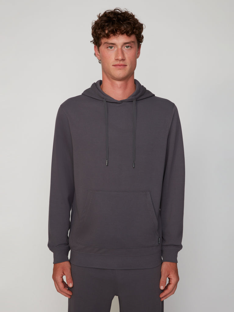 A young man with curly brown hair wears the TECH APPAREL MEN’S MICRO WAFFLE CLASSIC FIT POPOVER HOODIE in dark gray, featuring a front pocket and drawstrings, standing against a plain light gray background with a neutral expression.