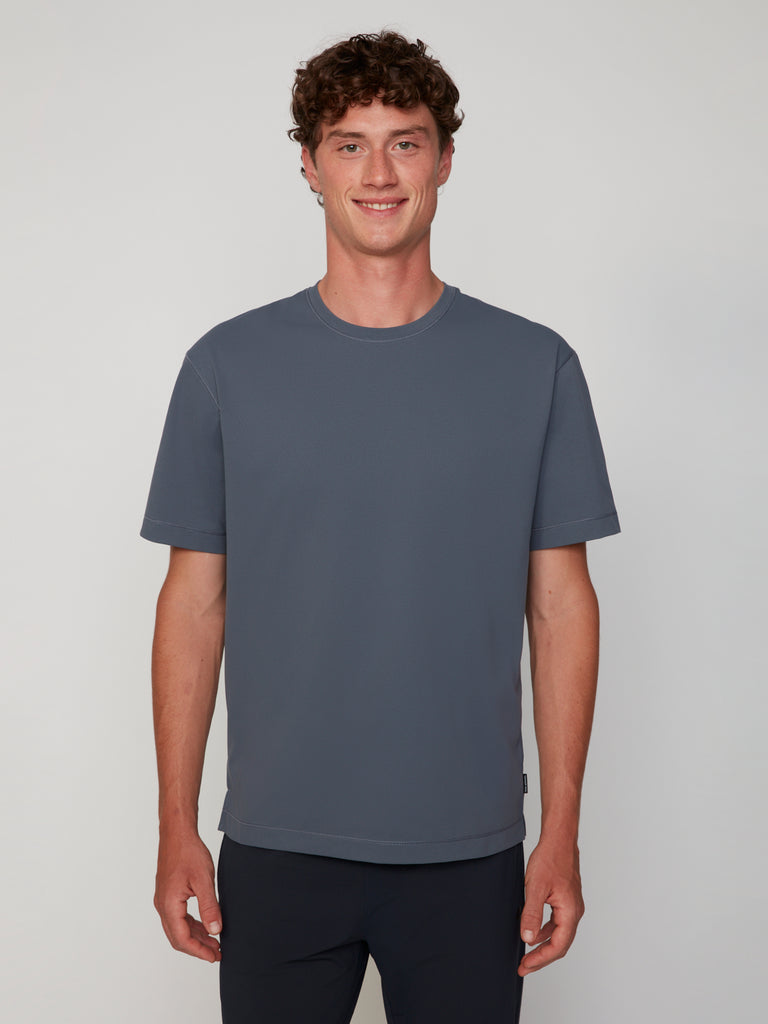 A young man with curly brown hair smiles while standing against a light gray background, wearing TECH APPAREL’s MEN’S PURE FEEL RELAXED DROP SHOULDER CREW NECK T-SHIRT in slate gray and dark pants.