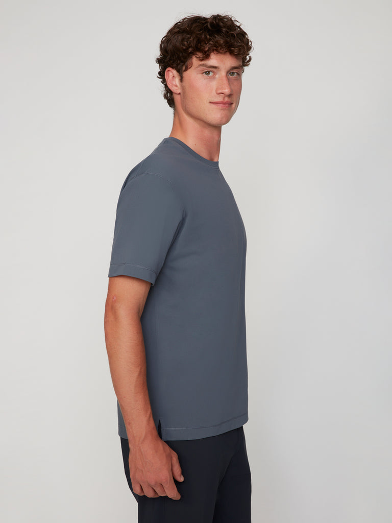A young man with curly brown hair stands sideways to the camera, wearing the TECH APPAREL MEN’S PURE FEEL RELAXED DROP SHOULDER CREW NECK T-SHIRT in gray and dark pants against a plain light background.