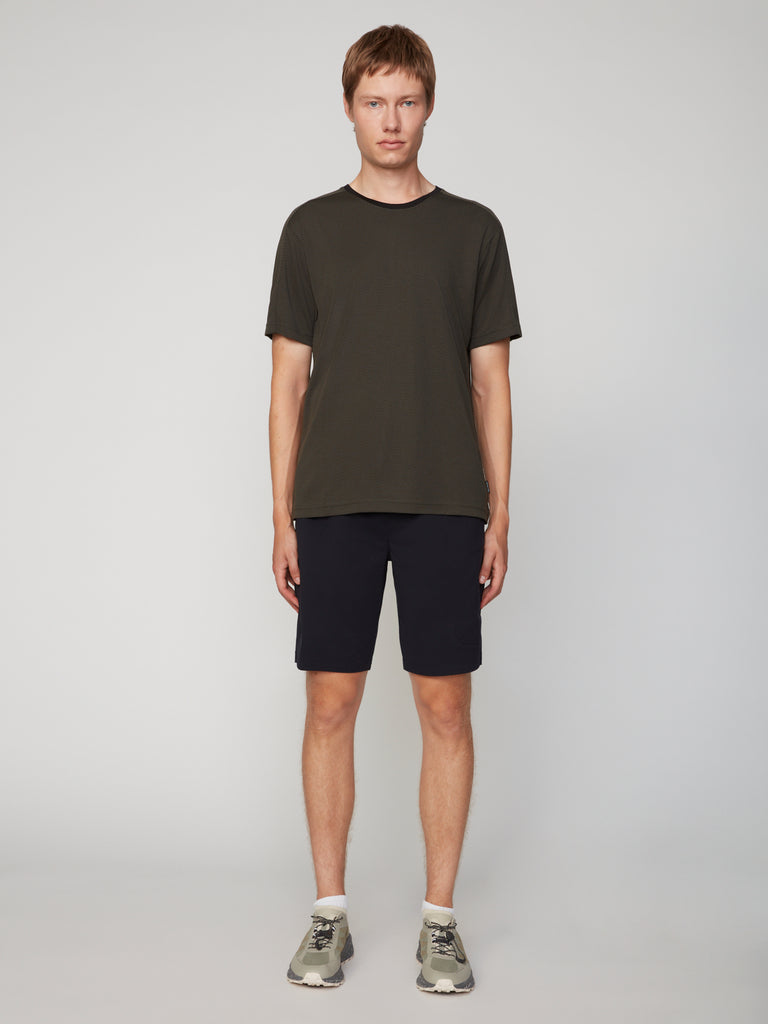 A young man stands facing forward against a light gray background, wearing a dark olive-green t-shirt and TECH APPAREL MEN’S 360:KNIT PULL-ON SHORT – 9.5 INSEAM, with white socks and gray sneakers. His arms rest naturally at his sides.