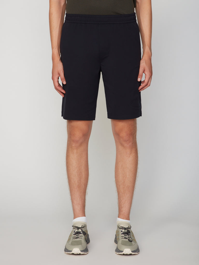 Only the lower half of a person is visible, showcasing TECH APPAREL MEN’S 360:KNIT PULL-ON SHORT – 9.5 INSEAM in a classic fit, paired with a dark t-shirt, white socks, and gray sneakers against a plain background.
