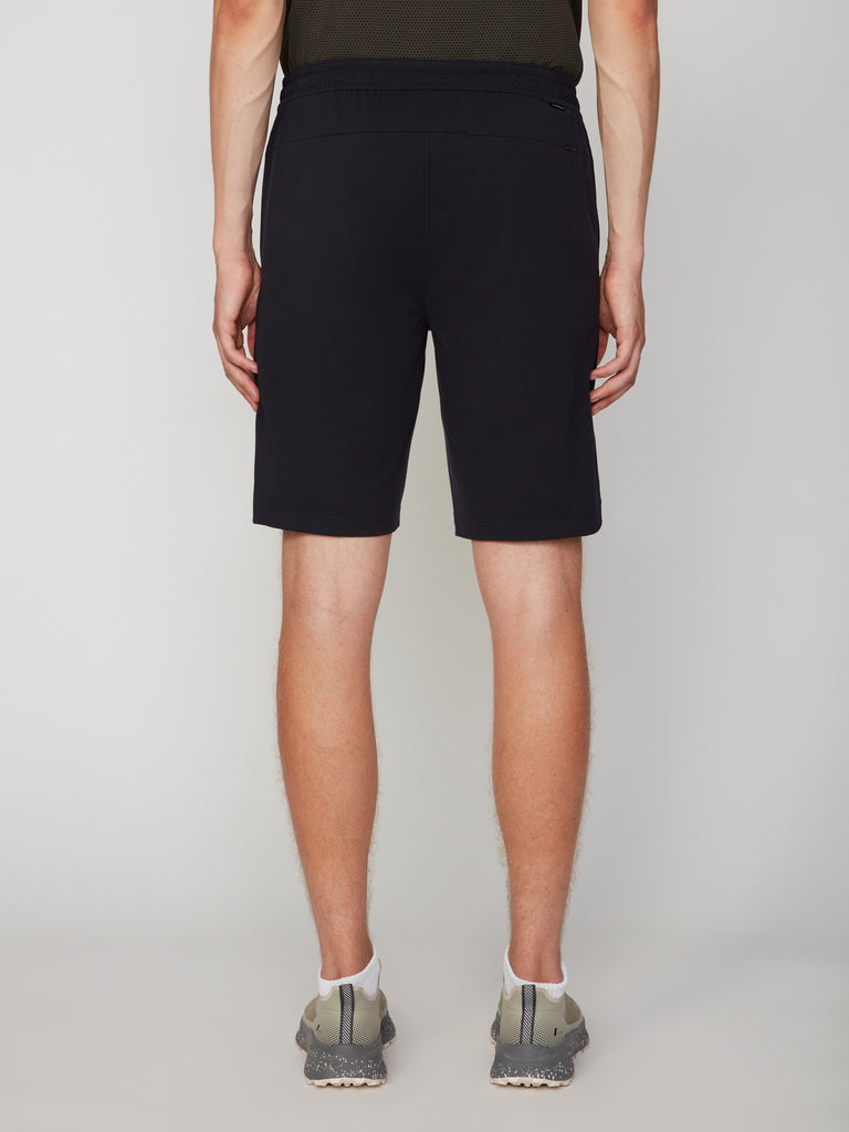 A person stands facing away, wearing a black shirt and TECH APPAREL MEN’S 360:KNIT PULL-ON SHORT – 9.5 INSEAM with stretch-engineered fabric. Gray sneakers complete the look against a plain, light background.