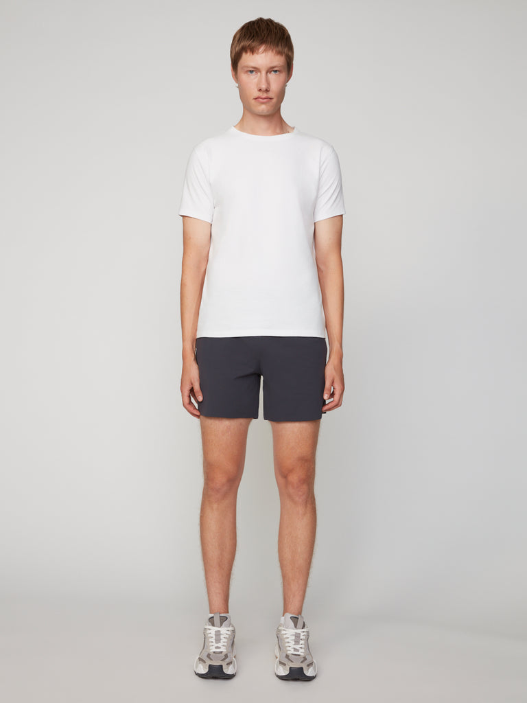 A person stands facing forward against a plain light background, wearing a white short-sleeve t-shirt, TECH APPAREL MEN’S 360:KNIT PULL-ON SHORT – 6 INSEAM in dark fabric, white socks, and gray sneakers.