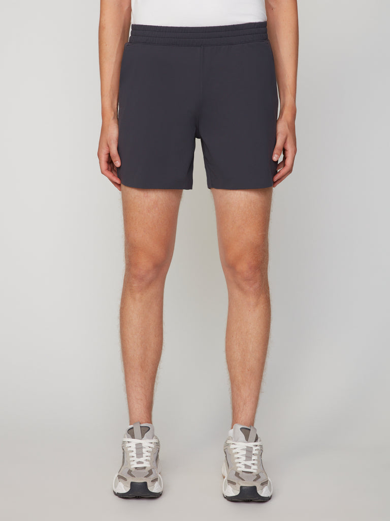 A person stands facing forward against a plain light background, wearing a white shirt, white sneakers, and TECH APPAREL MEN’S 360:KNIT PULL-ON SHORT – 6 INSEAM in black. The photo shows their body from shoulders to feet.