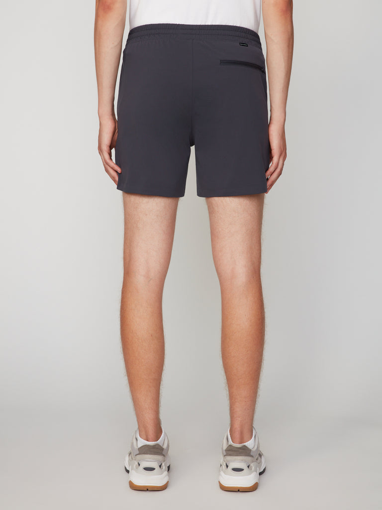 A person facing away from the camera wears a white shirt, TECH APPAREL MEN’S 360:KNIT PULL-ON SHORT – 6 INSEAM in dark gray, and white sneakers, set against a plain light background.