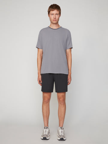 A person stands facing forward against a plain light background, wearing a gray t-shirt, TECH APPAREL MEN’S 360:FLEX PULL-ON SHORT – 7.5 INSEAM, and gray sneakers. Their arms are at their sides and their expression is neutral.