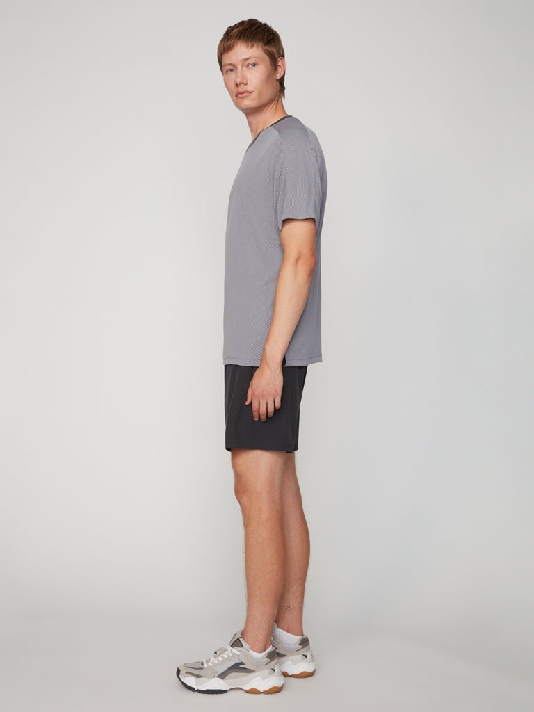 A person with short light brown hair stands sideways in a gray t-shirt and TECH APPAREL MEN’S 360:FLEX PULL-ON SHORT – 7.5 INSEAM, styled above the knee with white sneakers against a plain light gray background.
